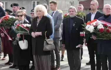 Uczestnicy uroczystości, niekt&oacute;rzy trzymają wiazanki kwiat&oacute;w.