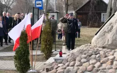 Uczestnicy uroczystości na tle flag biało czerwonych. Mundurowi salutują.