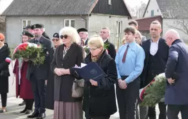 Uczestnicy uroczystości, niekt&oacute;rzy trzymają wiazanki kwiat&oacute;w.